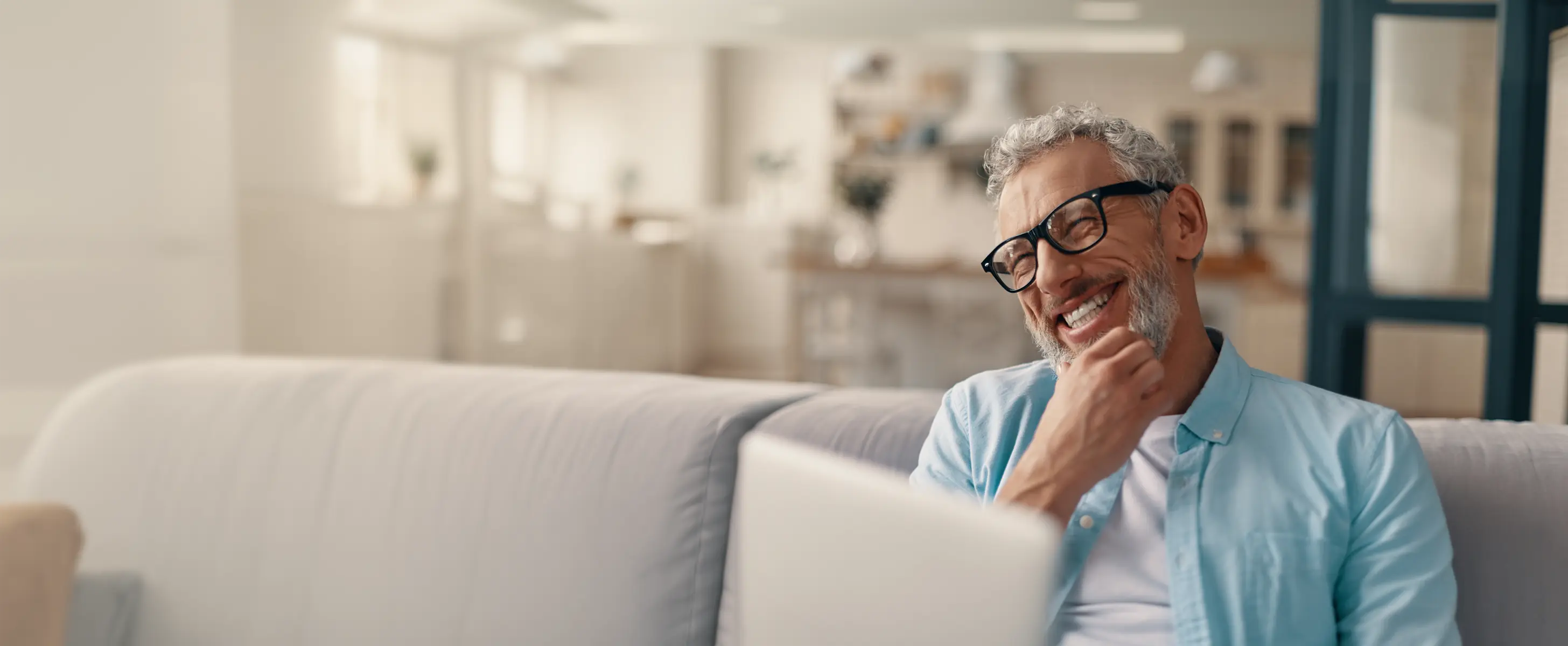 Smiling middle-aged man with glasses sitting on a couch, looking at a laptop.