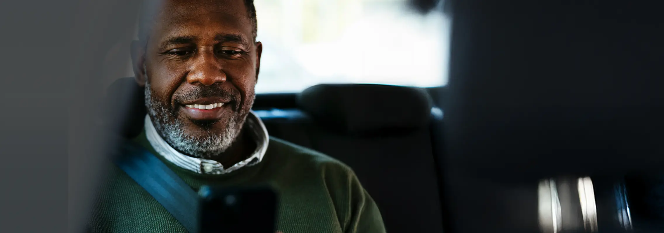 Smiling man wearing seatbelt sitting in a car and looking at his smartphone.
