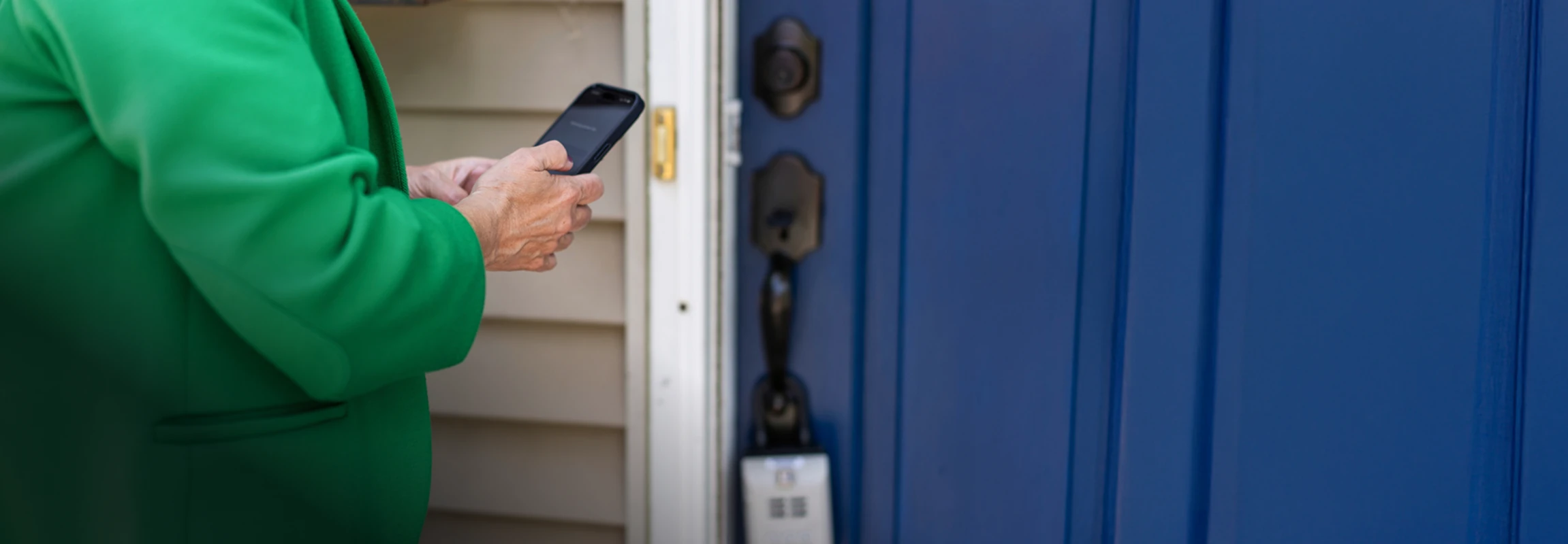 Person in green jacket holding a smartphone near a blue front door with a lock and doorbell.