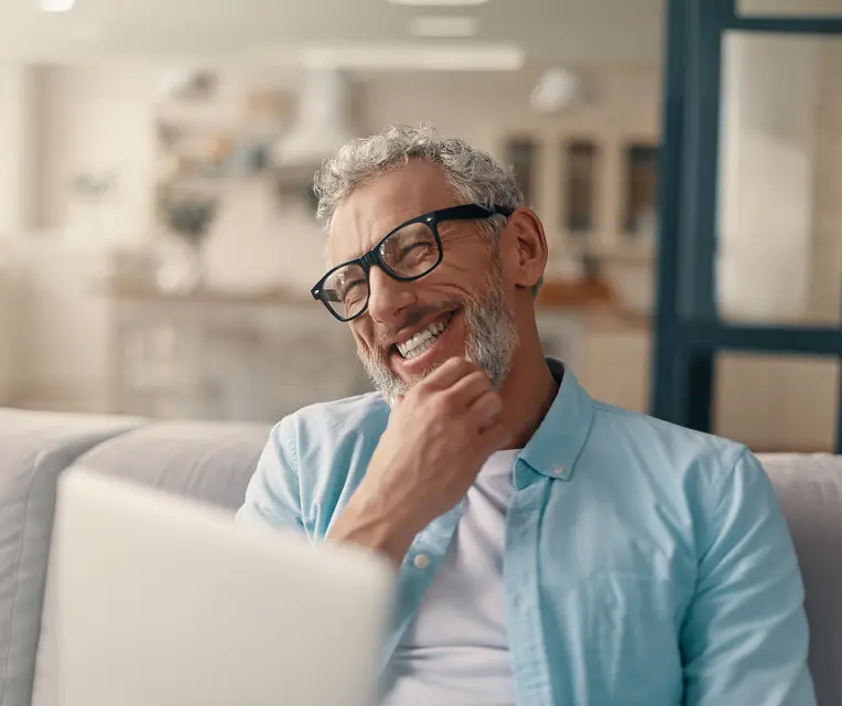 Smiling middle-aged man with gray hair and beard wearing glasses and a light blue shirt sitting on a couch.