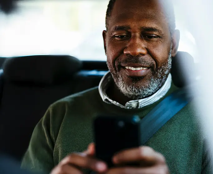 Smiling middle-aged man wearing a green sweater and seatbelt using a smartphone inside a car.