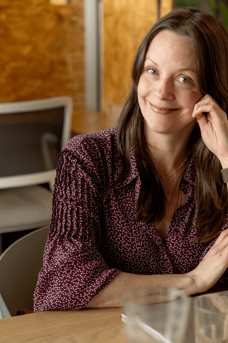 Sabrina, unsere Interface Designerin sitzt lächelnd an einem Holztisch in einem Innenraum und stützt den Kopf locker mit einer Hand ab. Sie trägt eine bordeauxrote Bluse mit kleinem weißen Punktmuster sowie eine Uhr am Handgelenk; im unscharfen Hintergrund sind Stühle und eine warme, holzfarbene Wand zu erkennen.