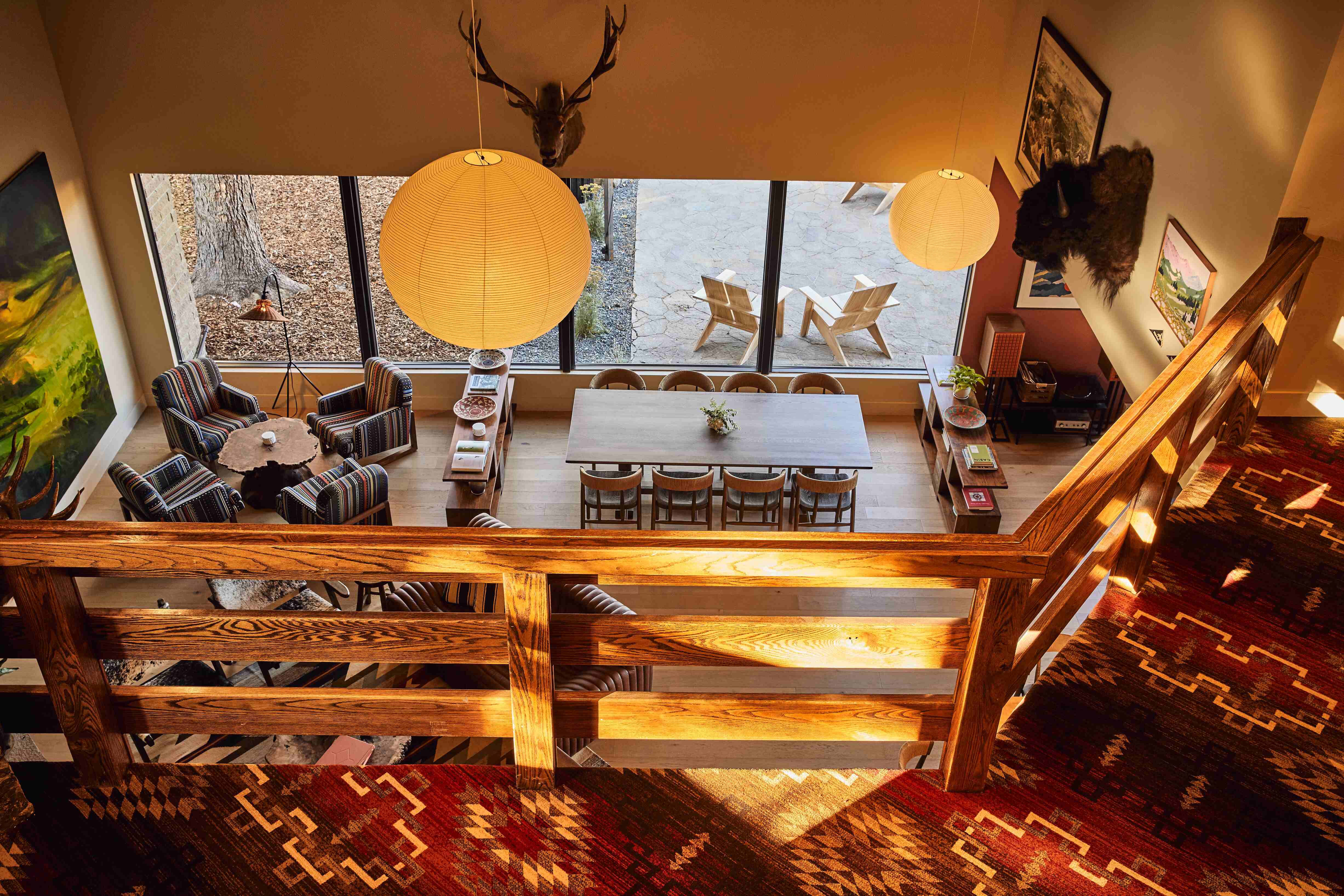 View from upstairs onto a cozy living and dining area with wooden furniture, patterned armchairs, large windows, pendant lamps, and mounted animal heads.