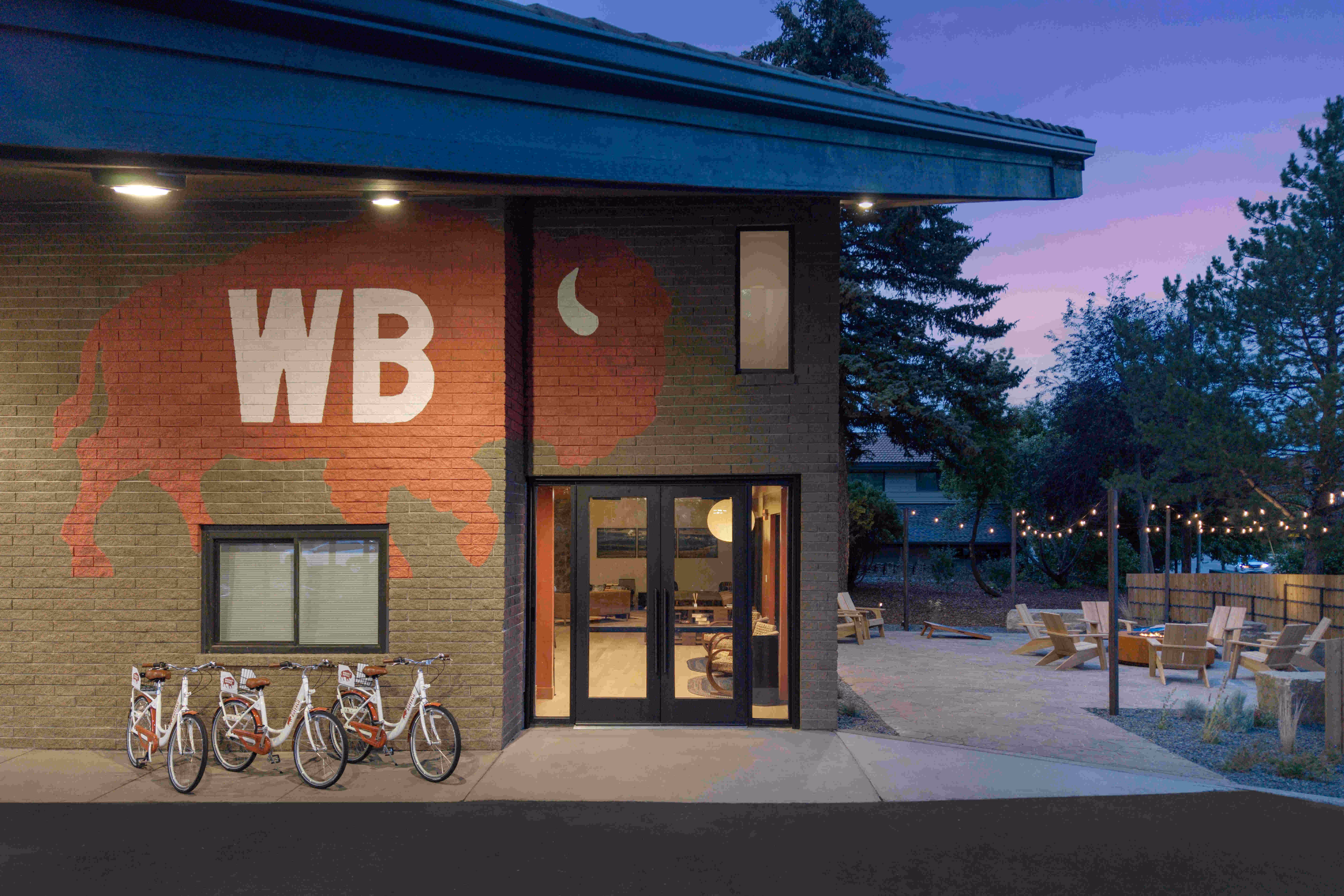 Building exterior at dusk with large red buffalo logo and white WB letters, four parked bicycles, and outdoor seating area with string lights.