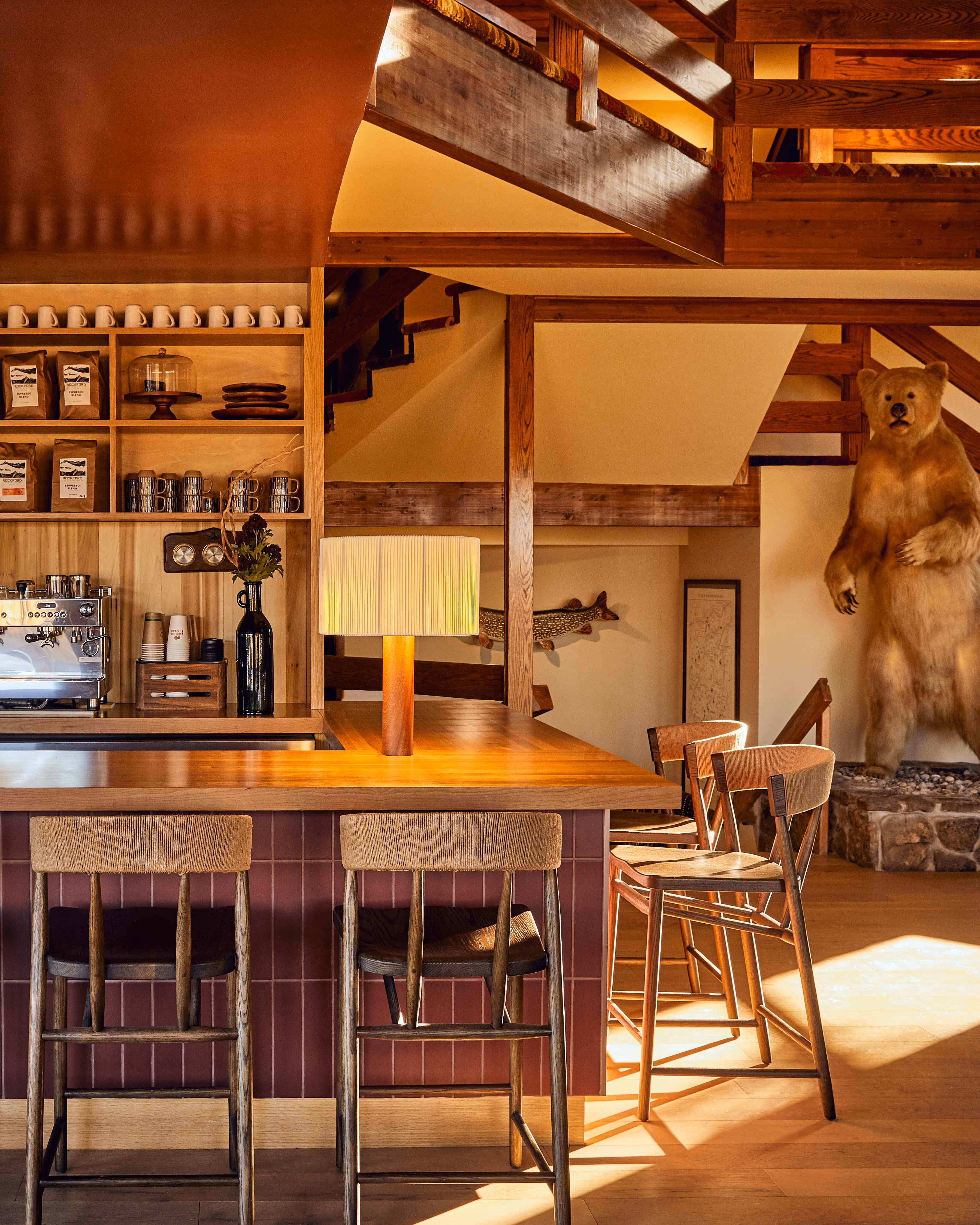 Cozy rustic bar area with wooden stools, a table lamp, coffee machine, shelves with mugs and coffee bags, and a taxidermy bear standing in the background.