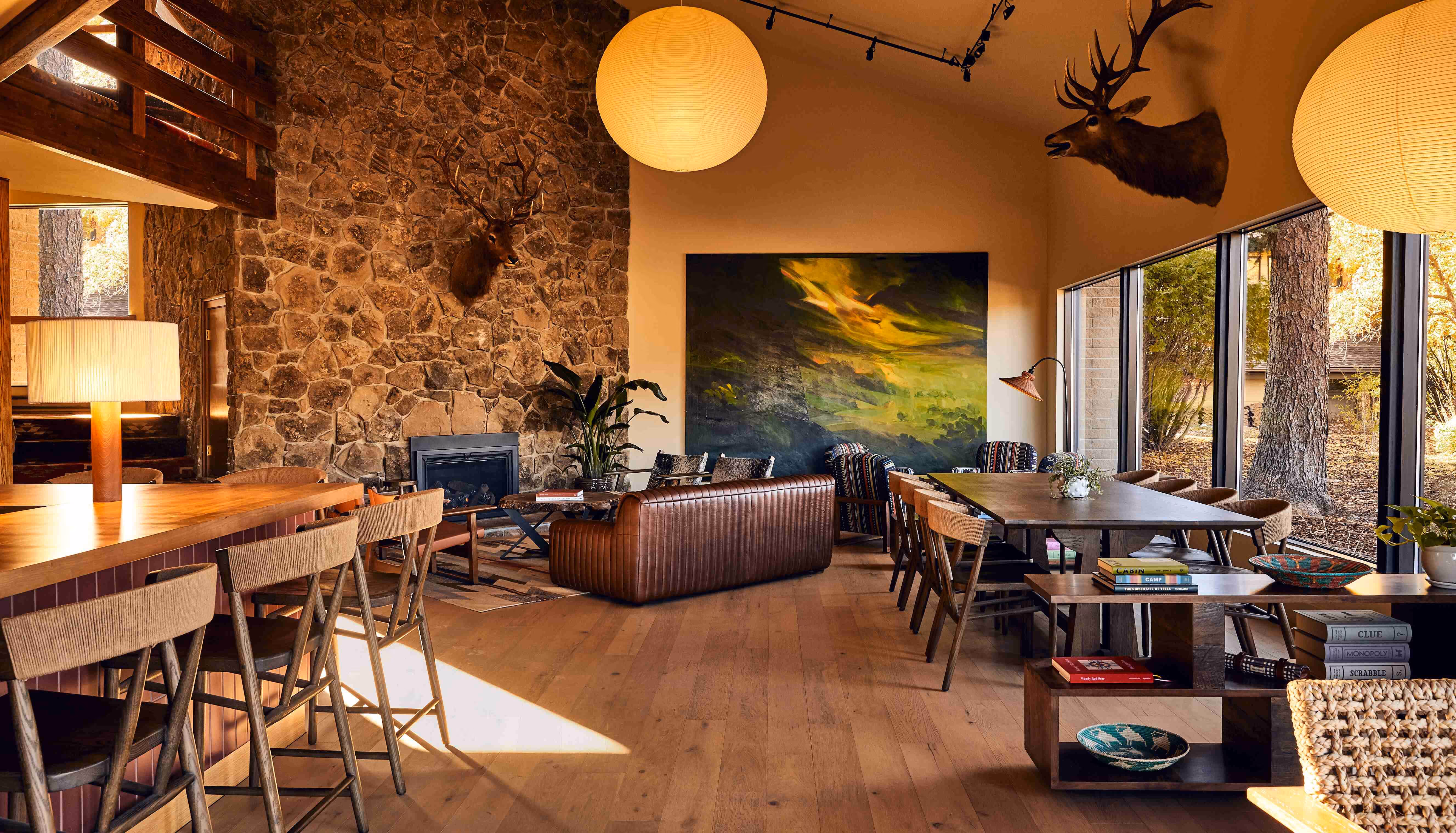 Cozy rustic living and dining area with stone fireplace, leather sofa, wooden chairs, large abstract painting, and mounted deer heads.
