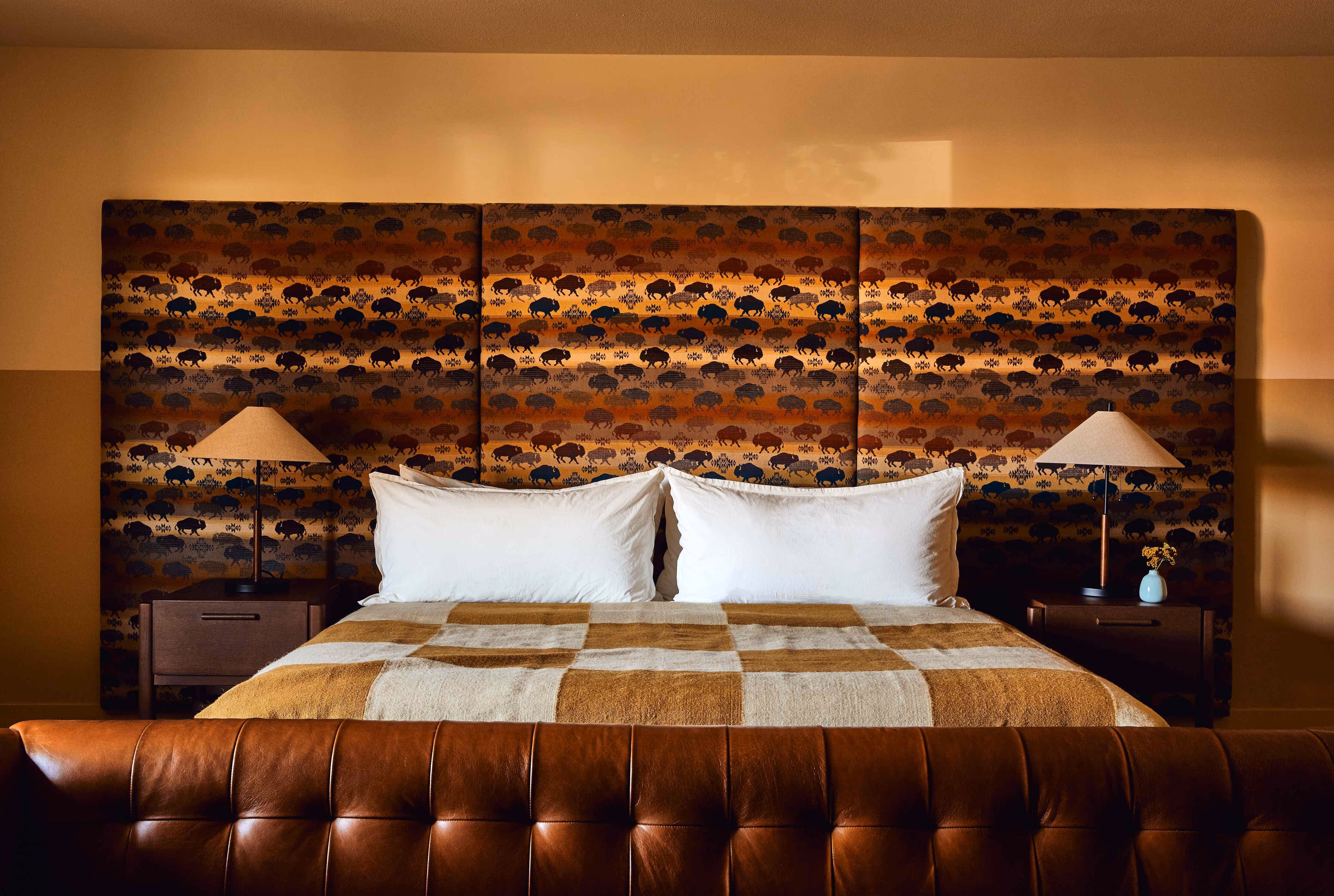 Bed with white pillows and a brown and beige checkered blanket, flanked by wooden nightstands with lamps and a patterned bison-themed headboard.
