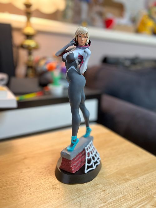 Painted figurine of Spider-Gwen standing in a dynamic pose atop a brick base with a white web detail, displayed on a wooden surface with a blurred background.