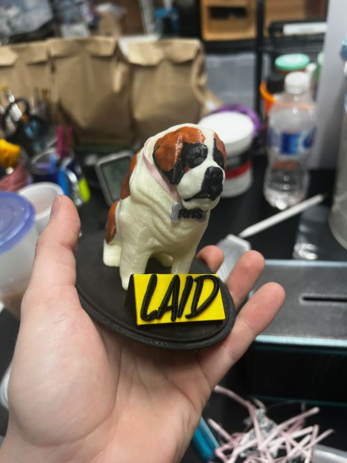 Hand holding a small figurine of a Saint Bernard dog wearing a pink collar with a “RHS” tag, seated on a black base with a bright yellow label that reads “LAID,” in front of a cluttered workshop background.