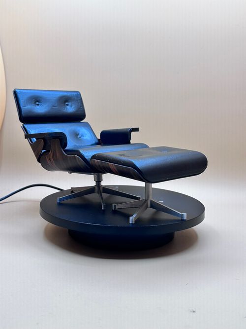 Miniature replica of a modern lounge chair and ottoman, styled like the Eames chair, set on a round black platform against a plain backdrop.