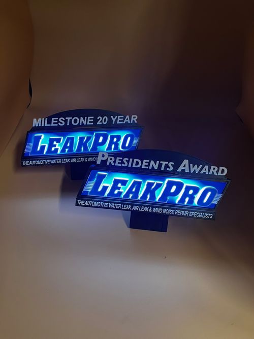 Two illuminated blue 3d printed award signs for “LeakPro,” one labeled “Milestone 20 Year” and the other “Presidents Award,” glowing against a neutral curved backdrop.
