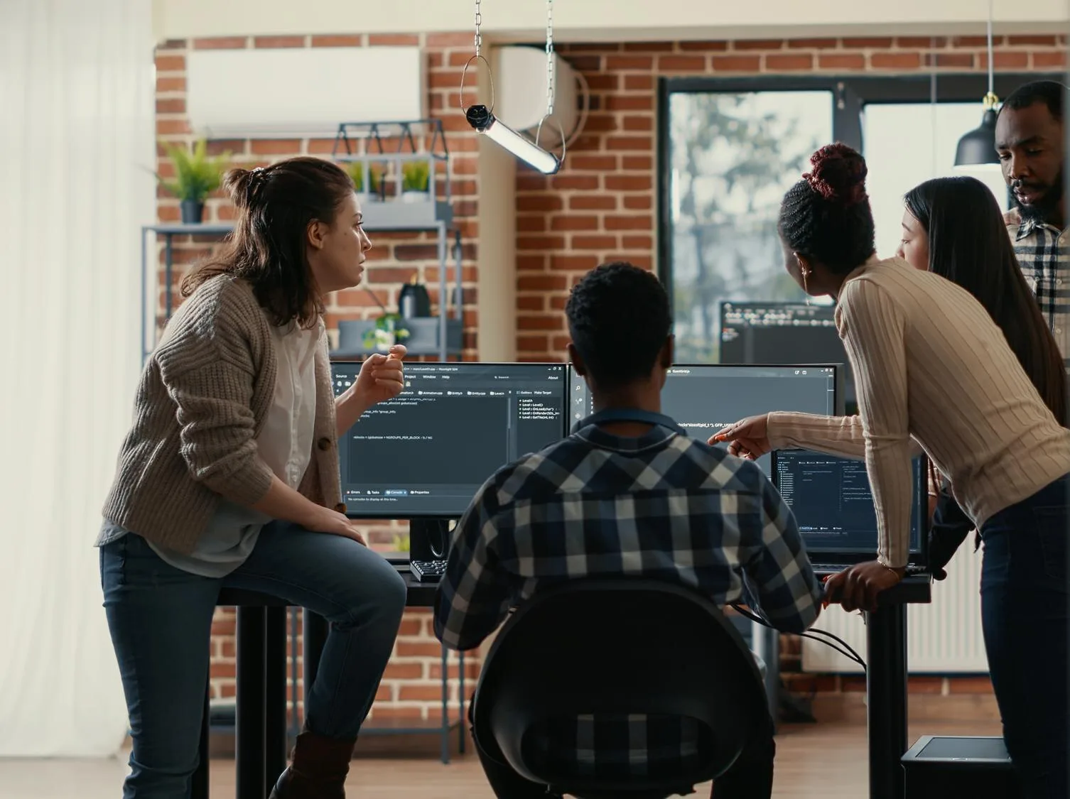 Five diverse coworkers collaborating and discussing computer code displayed on multiple monitors in a modern office.