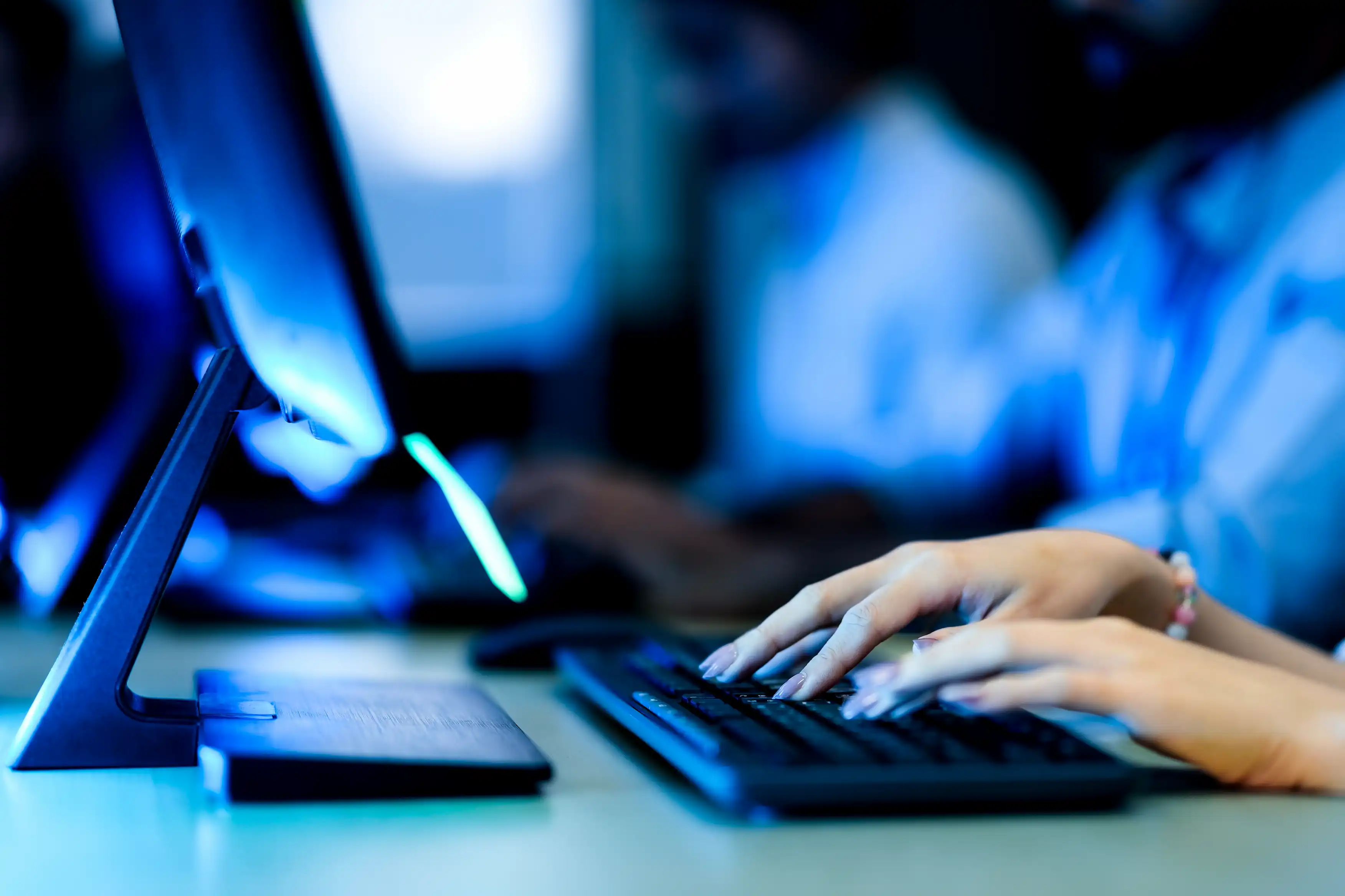 Close-up of hands typing on a keyboard with a computer monitor on a desk in a dimly lit setting.