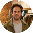 Man with curly dark hair wearing a beige jacket and gray shirt, standing indoors with clothes hanging in the background.