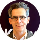 Portrait of a middle-aged man with gray hair and glasses smiling against a dark background.