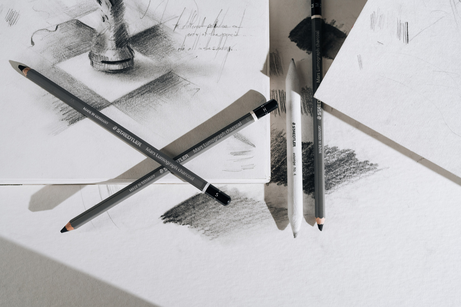 Four charcoal pencils and a blending stump on white paper with charcoal sketches and shading tests.