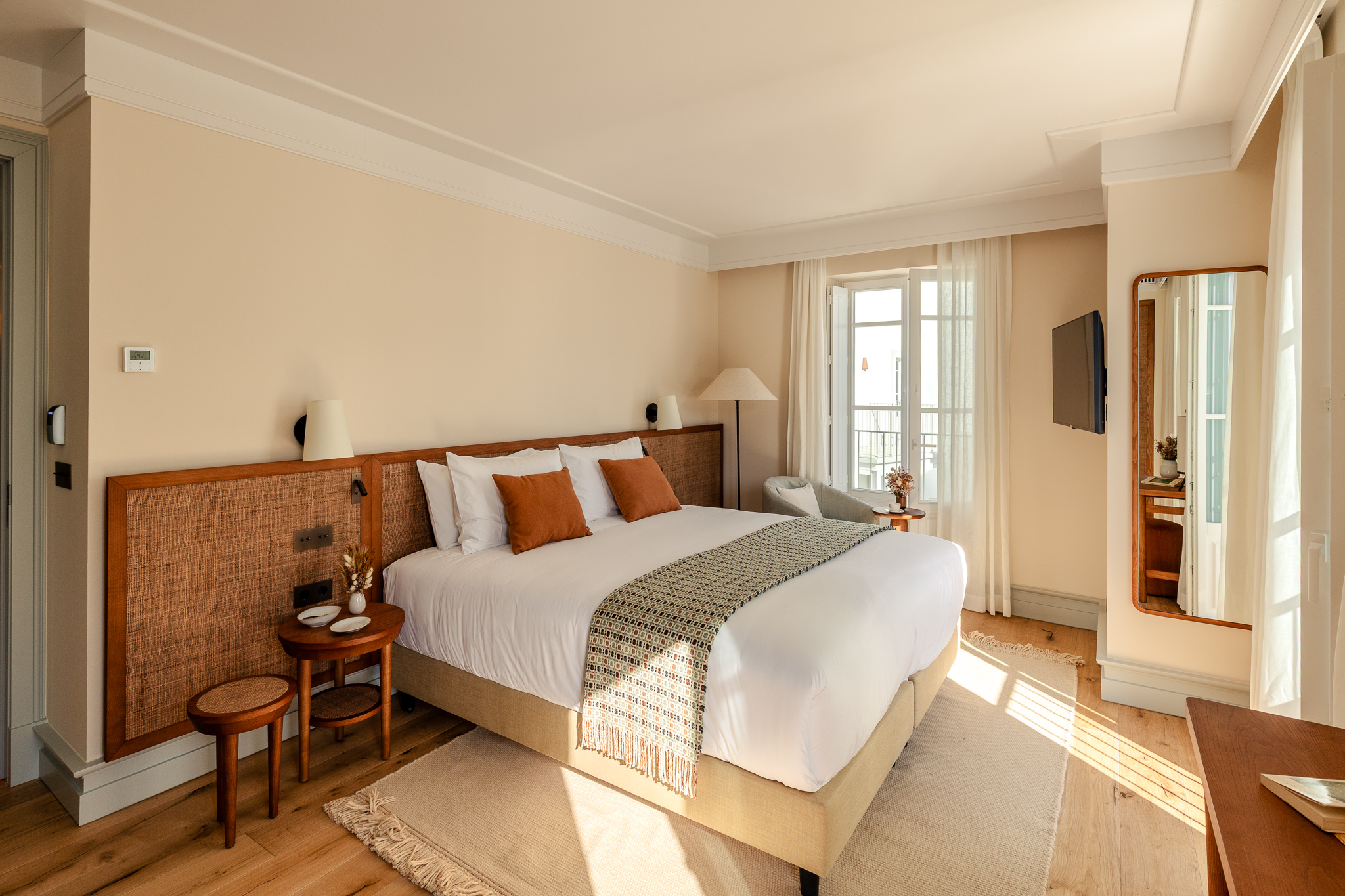 Charming hotel suite with a large double bed, gray pillows, patterned throw, soft lighting, and a small sofa at the foot of the bed holding a straw bag and red scarf - Palacio de Tavira