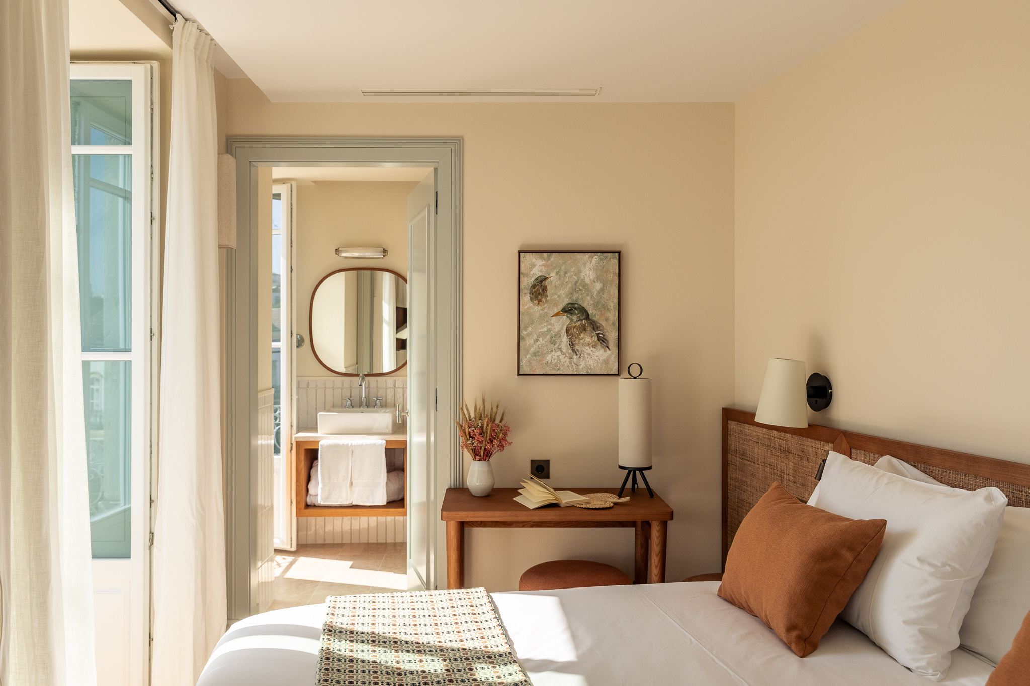 Stylish hotel corner with a wooden desk, a large ceramic lamp, an upholstered blue chair with a straw tote and red scarf, and a tall mirror reflecting the calm decor - Palacio de Tavira