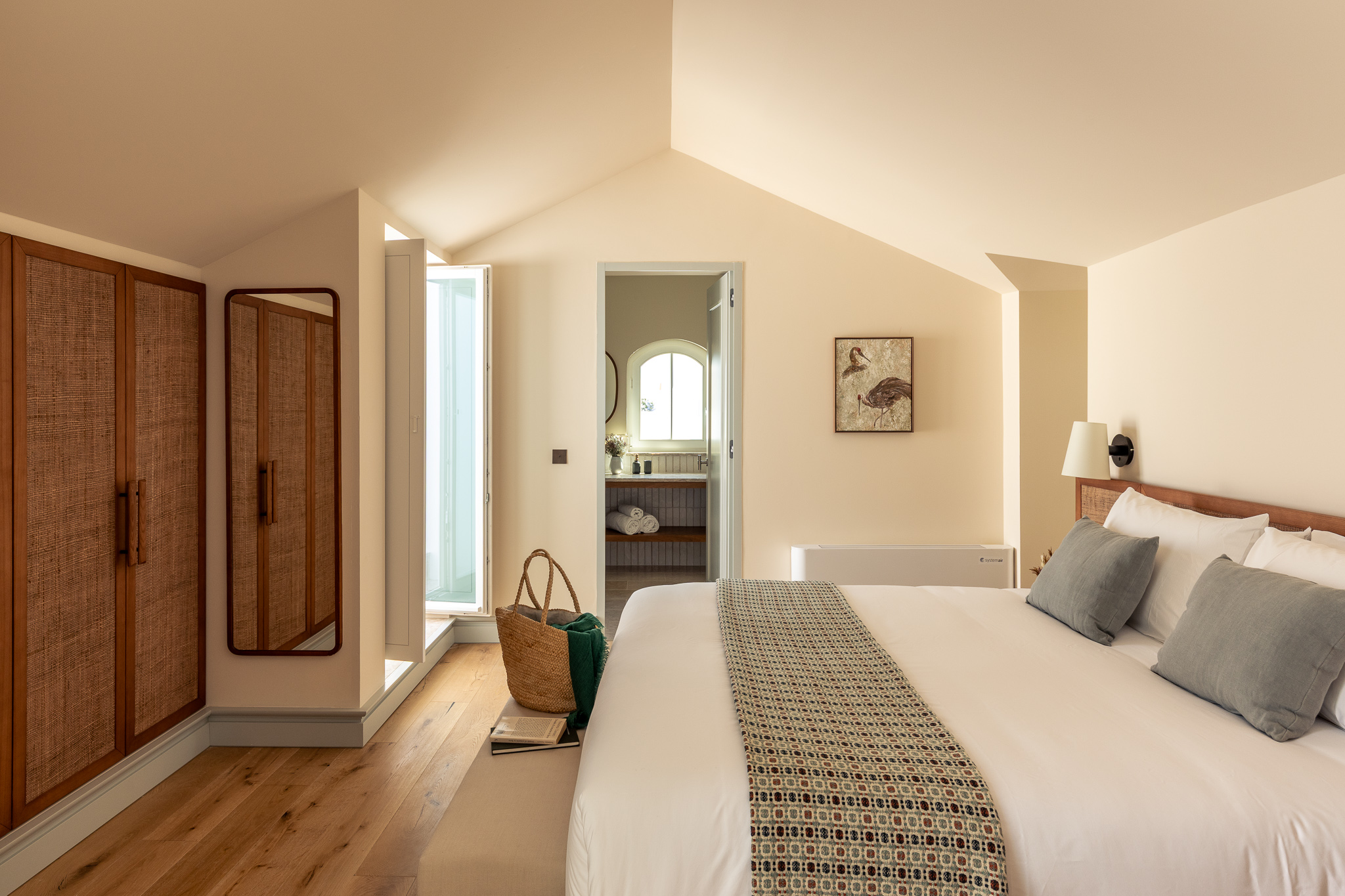 Warm and inviting hotel bed setup with a gray pillow arrangement, a colorful bedspread, and bedside table featuring fruit, water, and books under a glowing wall lamp - Palacio de Tavira