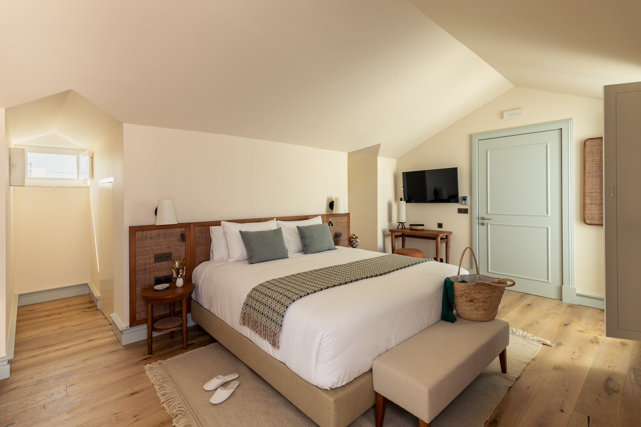 Bright and cozy junior suite attic room with minimalist decor, a well-dressed bed with orange and white accents, and natural wood furniture beside large curtained windows - Palacio de Tavira