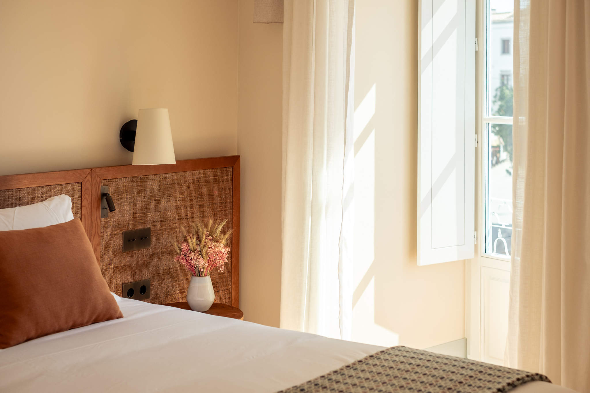 Close-up of a cozy hotel bed with a light gray cushion, white linens, and a patterned throw, next to a wooden side table with a vase of fresh yellow and red flowers under a wall lamp - Palacio de Tavira
