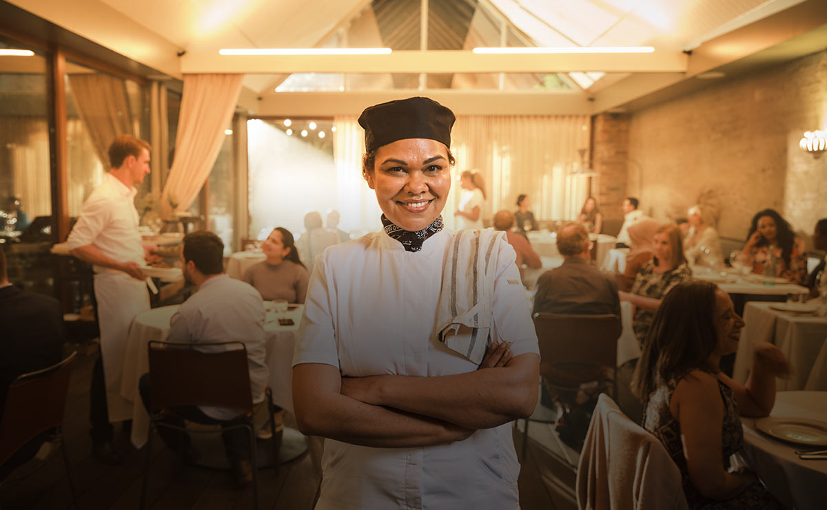 A hospitality worker smiles at the camera in front of a busy restaurant setting.