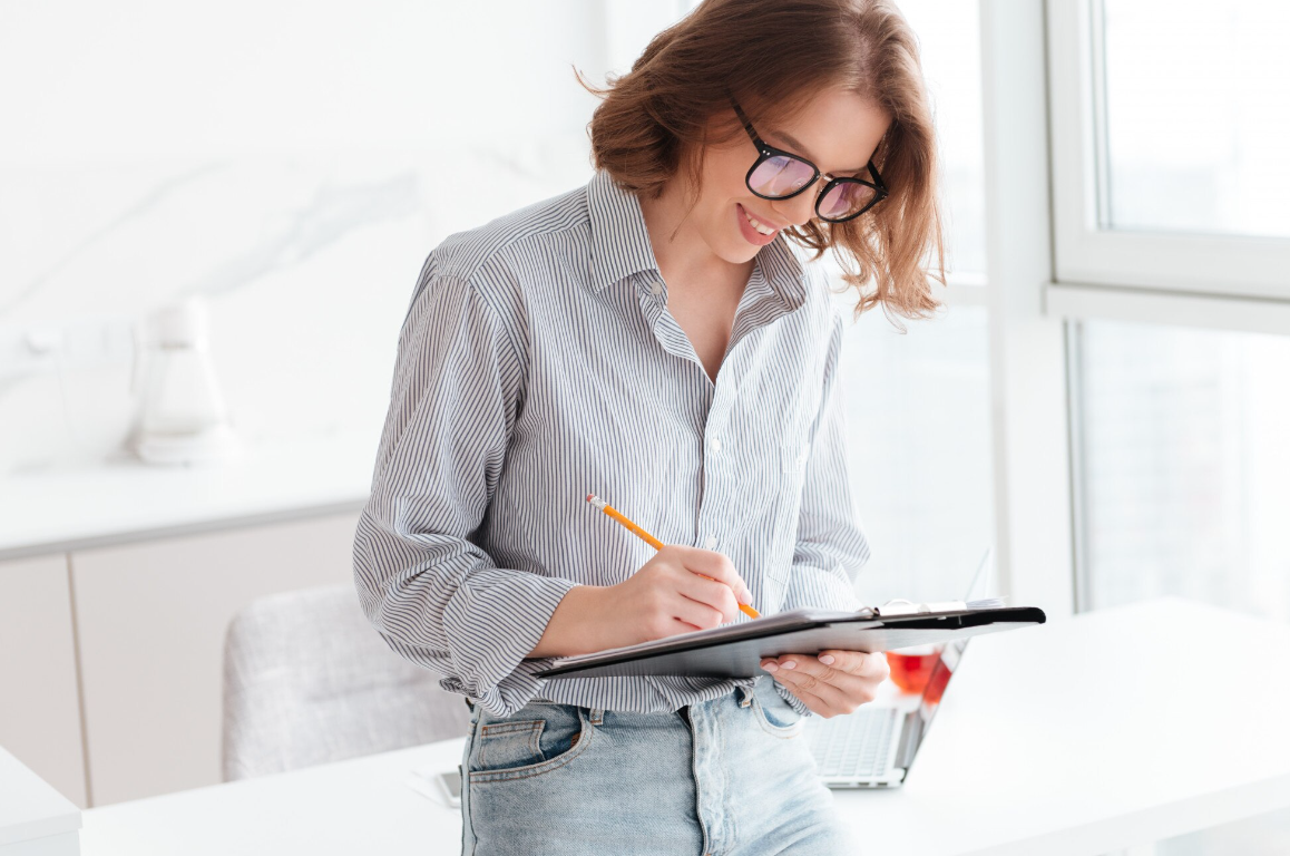 A smiling woman stands near a bright window, taking notes on a clipboard, representing progress tracking and personalized treatment planning in inpatient recovery