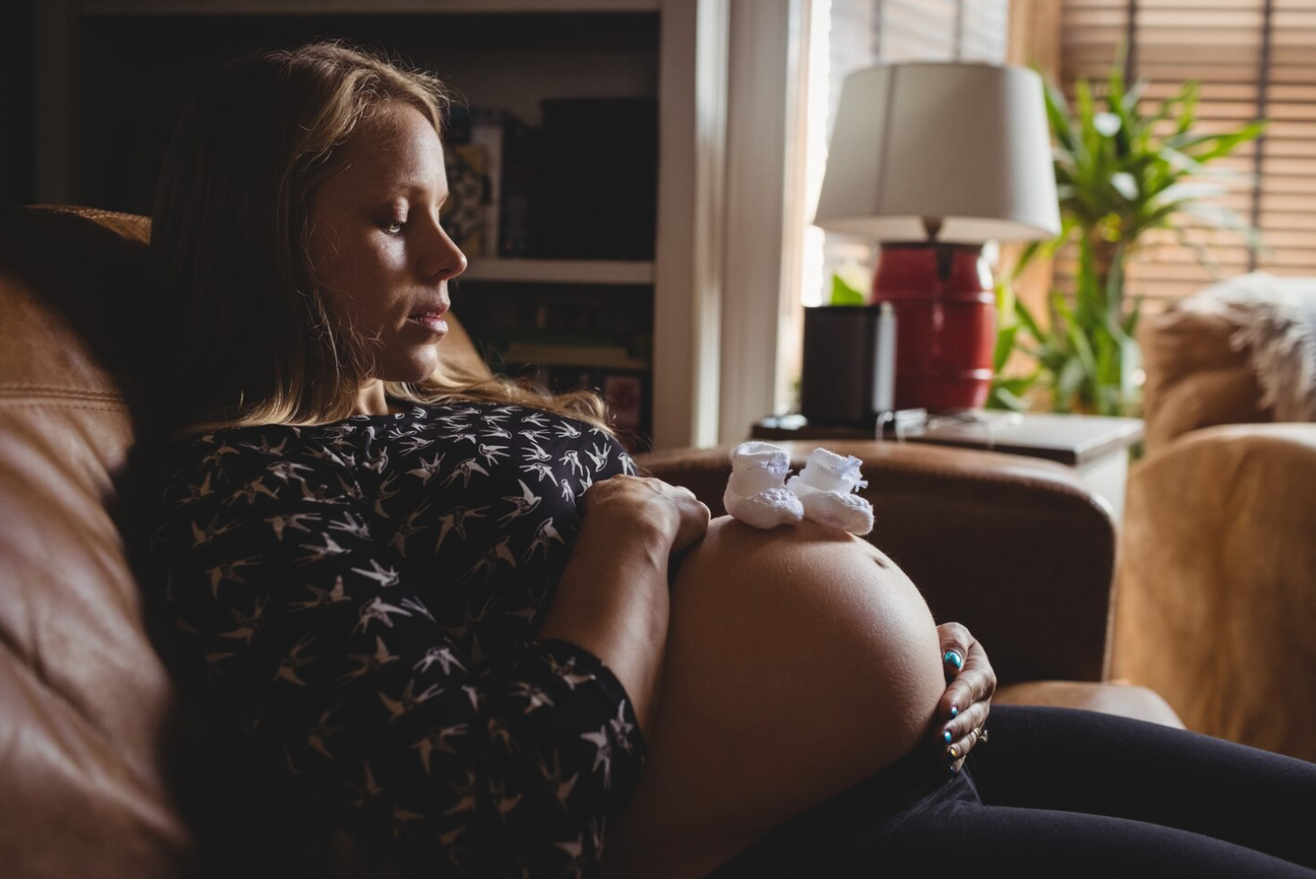 A pregnant woman sitting on a couch, looking at her womb and the pair of little shoes that rest on top of it
