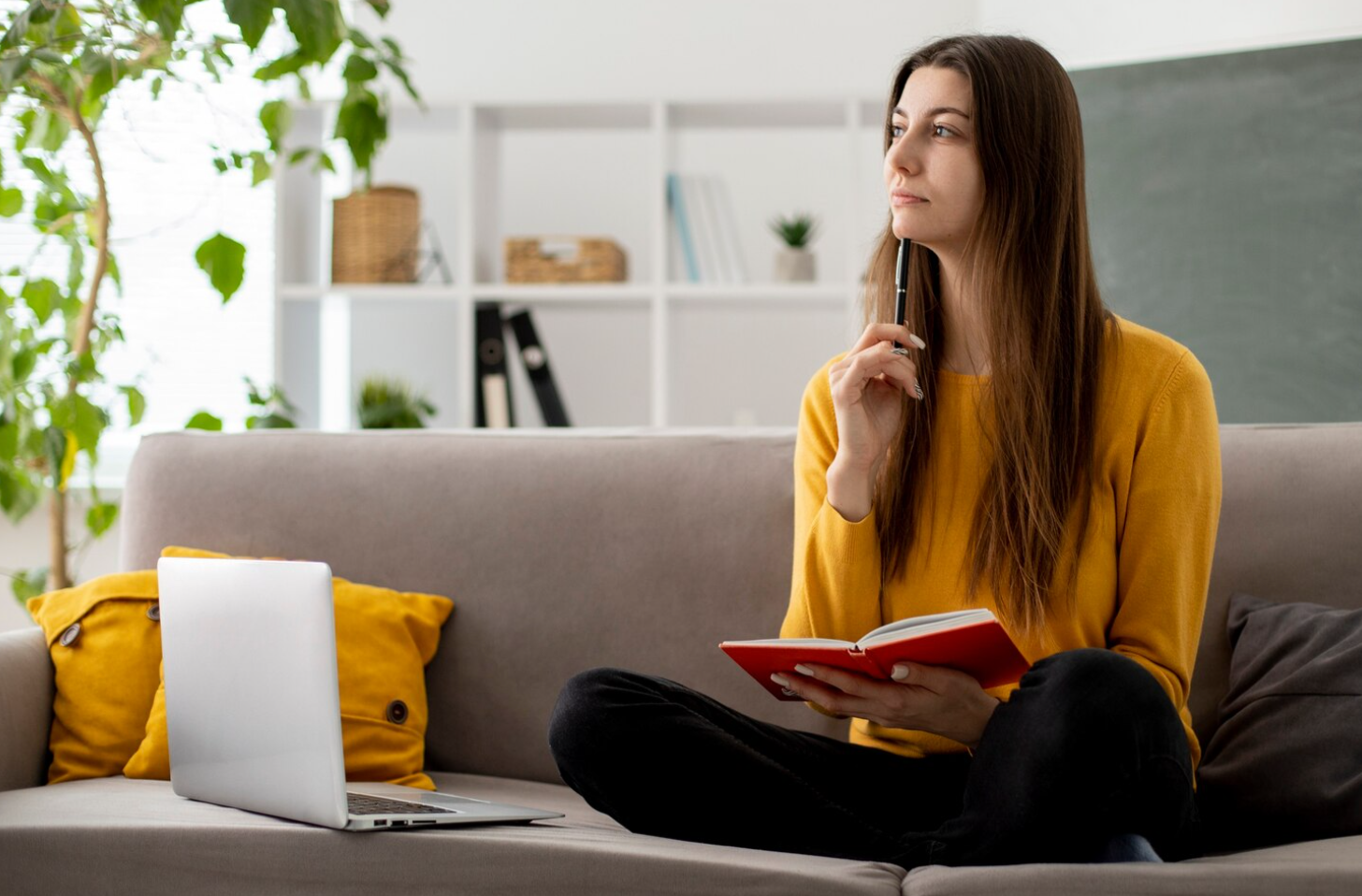 A woman sitting on a couch holding a notebook and a pen while thinking