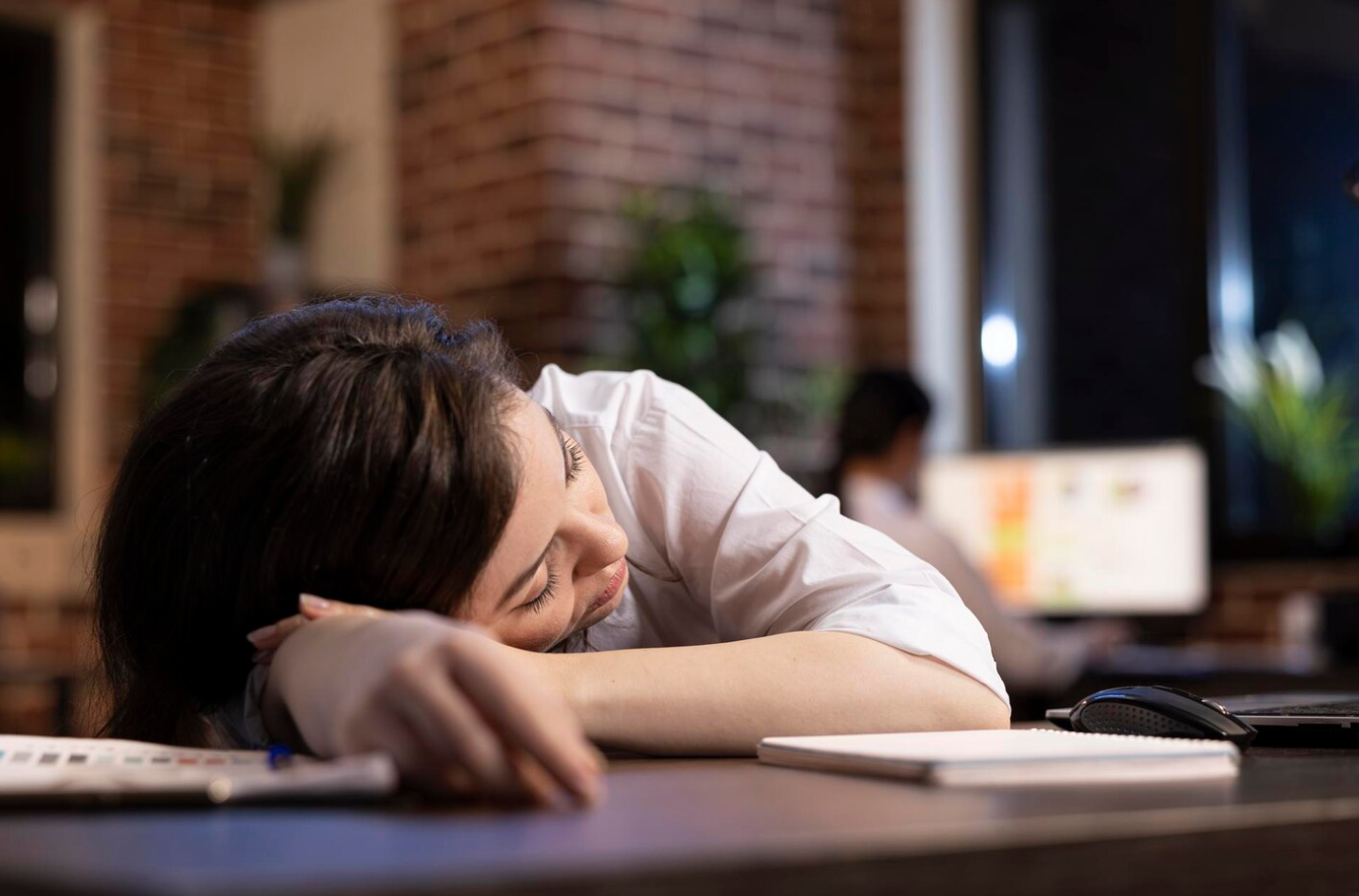 A woman is sleeping on a desk with her head resting on her arms, in front of a computer screen, demonstrating fatigue after quitting alcohol