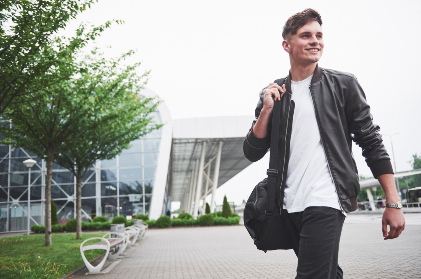 Young man in a leather jacket walking confidently outside a modern building with a duffel bag, representing independence and transition into sober living