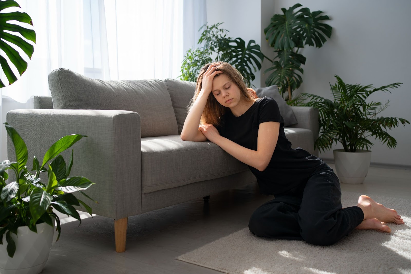 A woman kneels beside a sofa in a sunlit room, appearing fatigued and reflective, representing emotional exhaustion during meth withdrawal