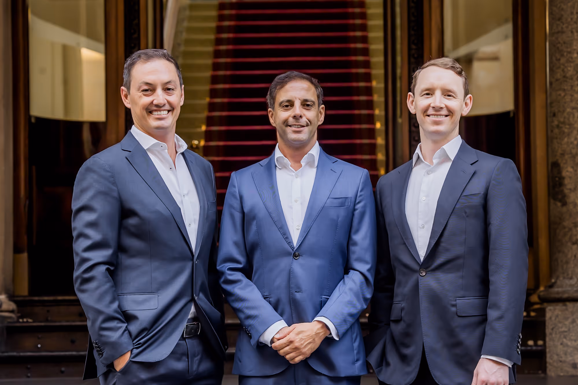 Three men in suits smiling and standing in front of a red-carpeted staircase in a formal setting.