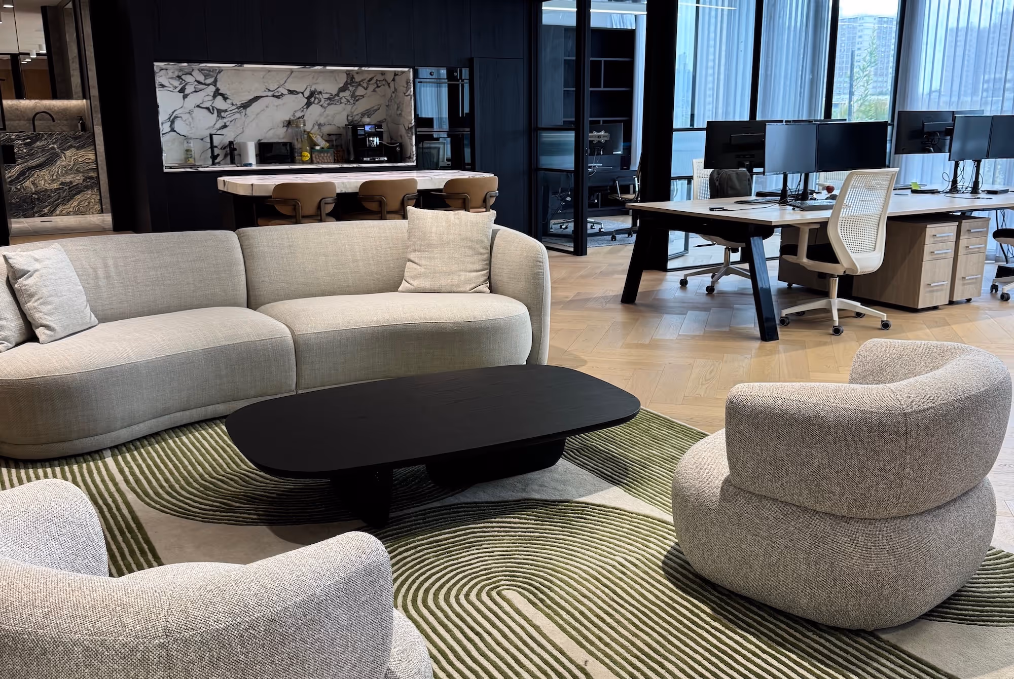 Modern office lounge area with a curved beige sofa, two rounded armchairs, a black oval coffee table, and a green patterned rug, next to workstations and a kitchen with marble backsplash.