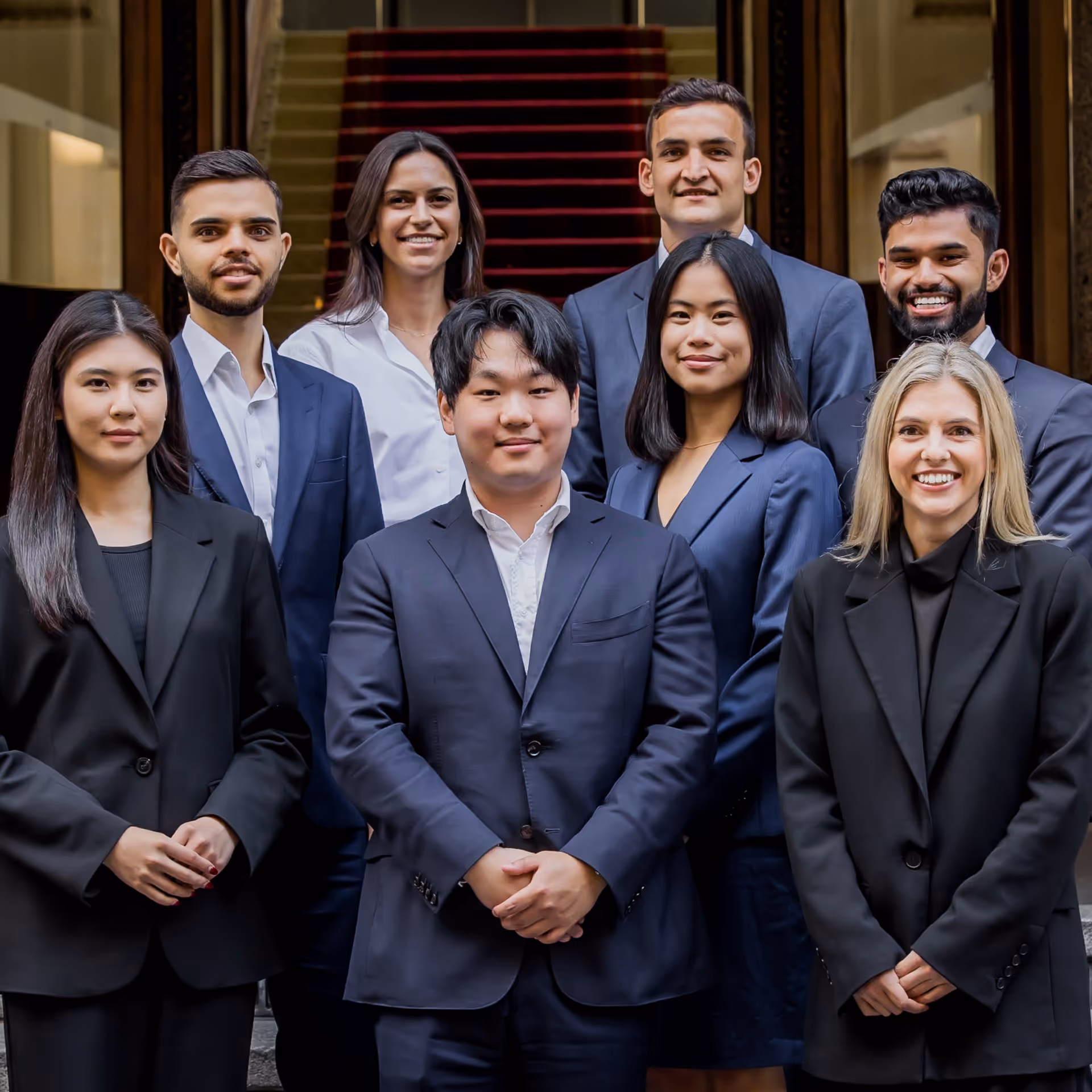A diverse group of eight young professionals dressed in business attire standing and smiling on stairs.