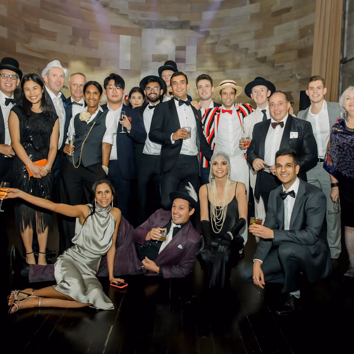 Group of well-dressed people at a party, many wearing black-tie outfits and vintage-style hats, some holding drinks and smiling.