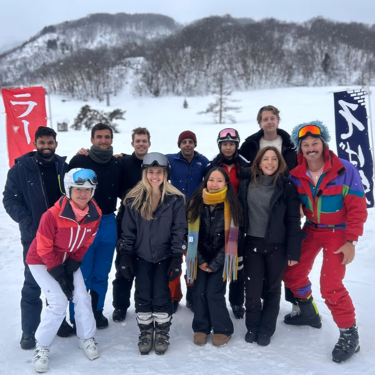 Group of ten people dressed in winter and ski clothing posing together on snow with snowy mountains in the background.