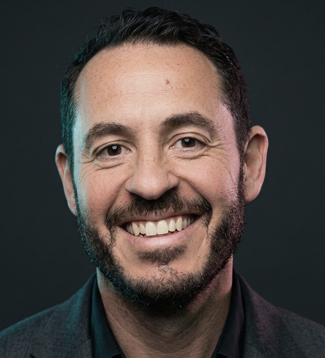 Smiling man with short dark hair, a trimmed beard, and wearing a dark blazer against a dark background.