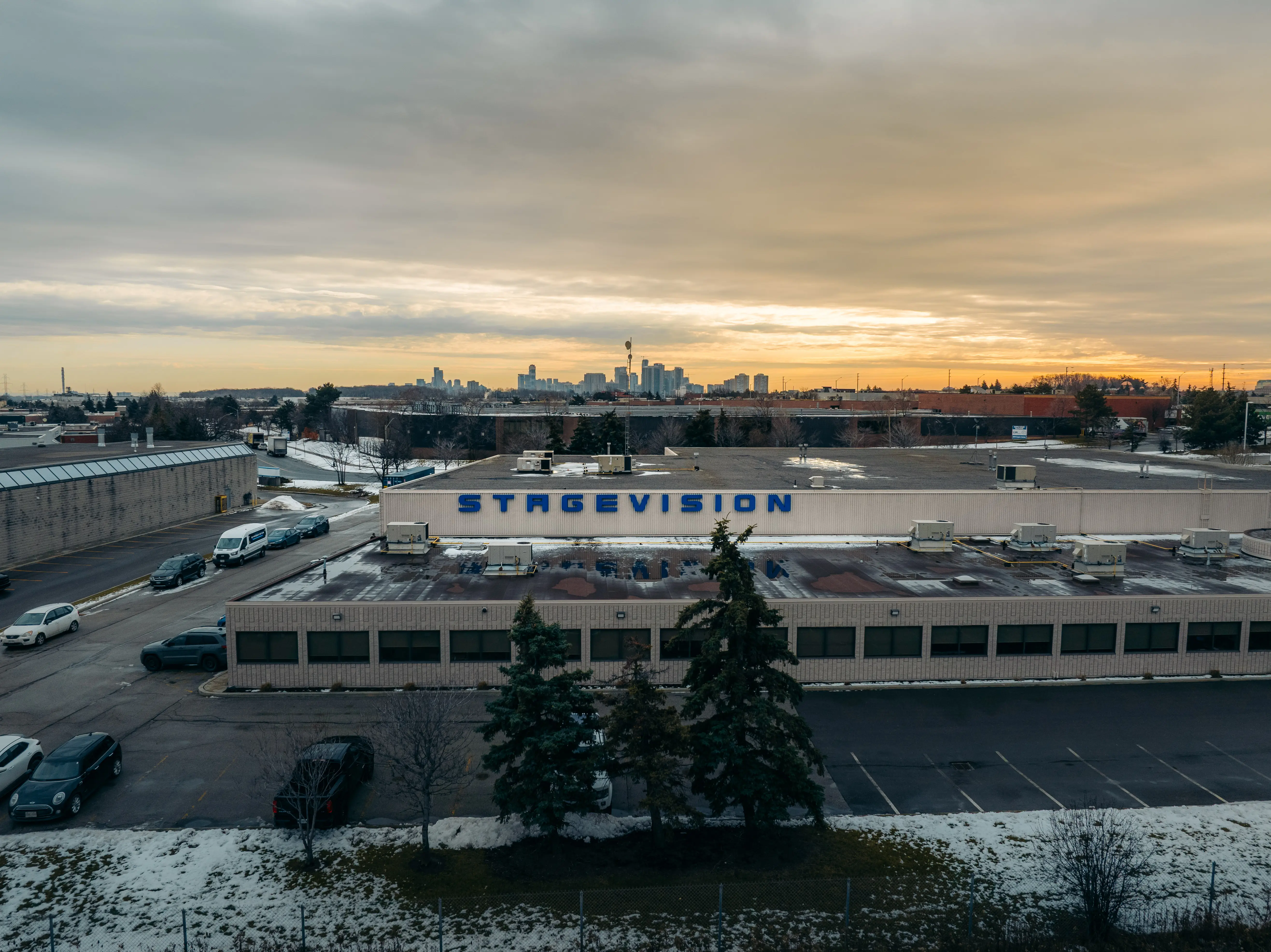 Industrial building with STAGEVISION sign on roof, parked cars, snowy foreground, and a city skyline under a cloudy sunset sky.