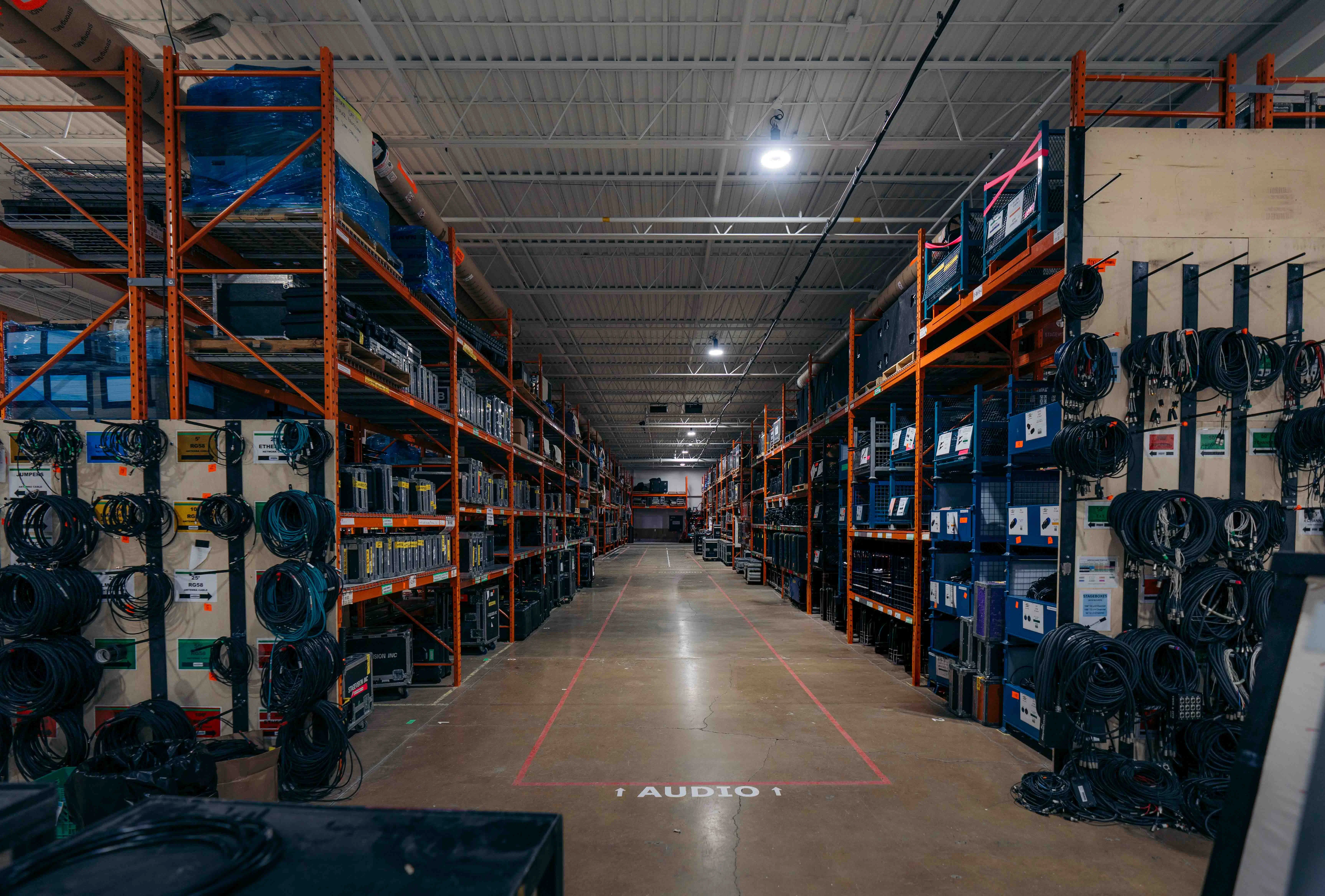 Warehouse aisle with shelves stocked with cables, audio equipment, and storage cases under overhead lighting.