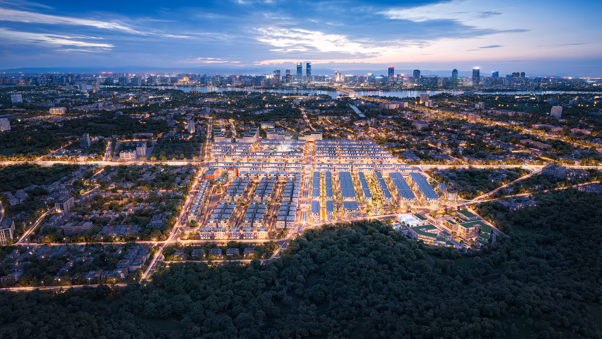 Aerial view of a large, illuminated urban residential area at dusk with a dense forest in the foreground and a city skyline across a river in the background.