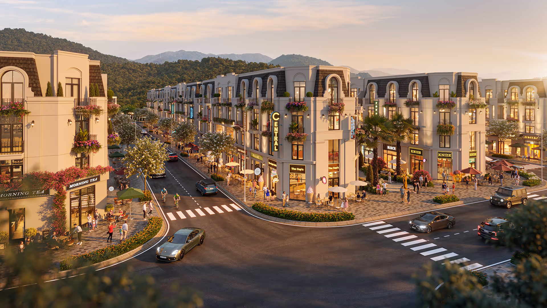 Elegant town square with multiple retail shops and cafes, people walking and sitting outdoors, cars on the road, surrounded by greenery and mountains at sunset.