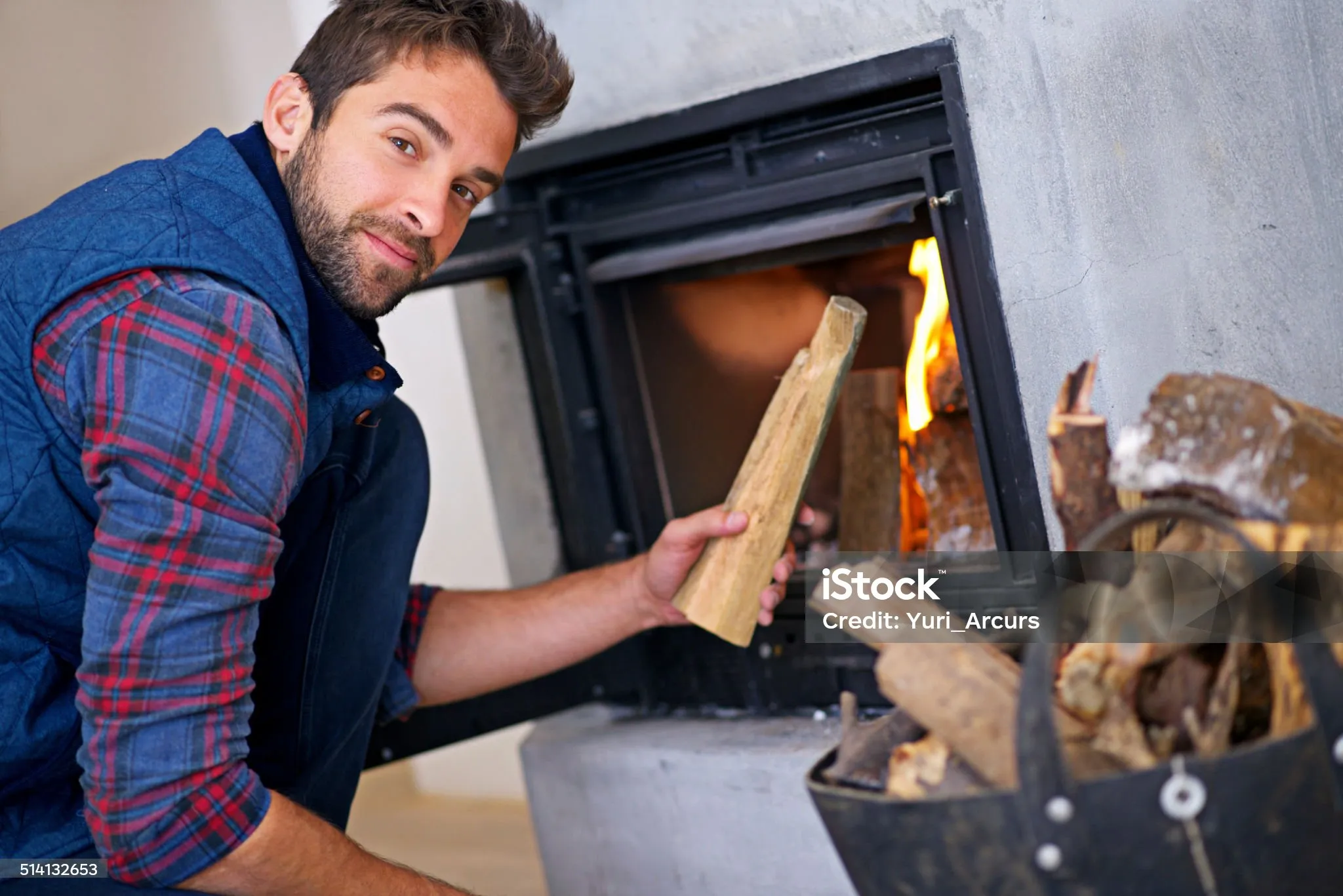 Man in plaid shirt adding wood to a burning fireplace at home
