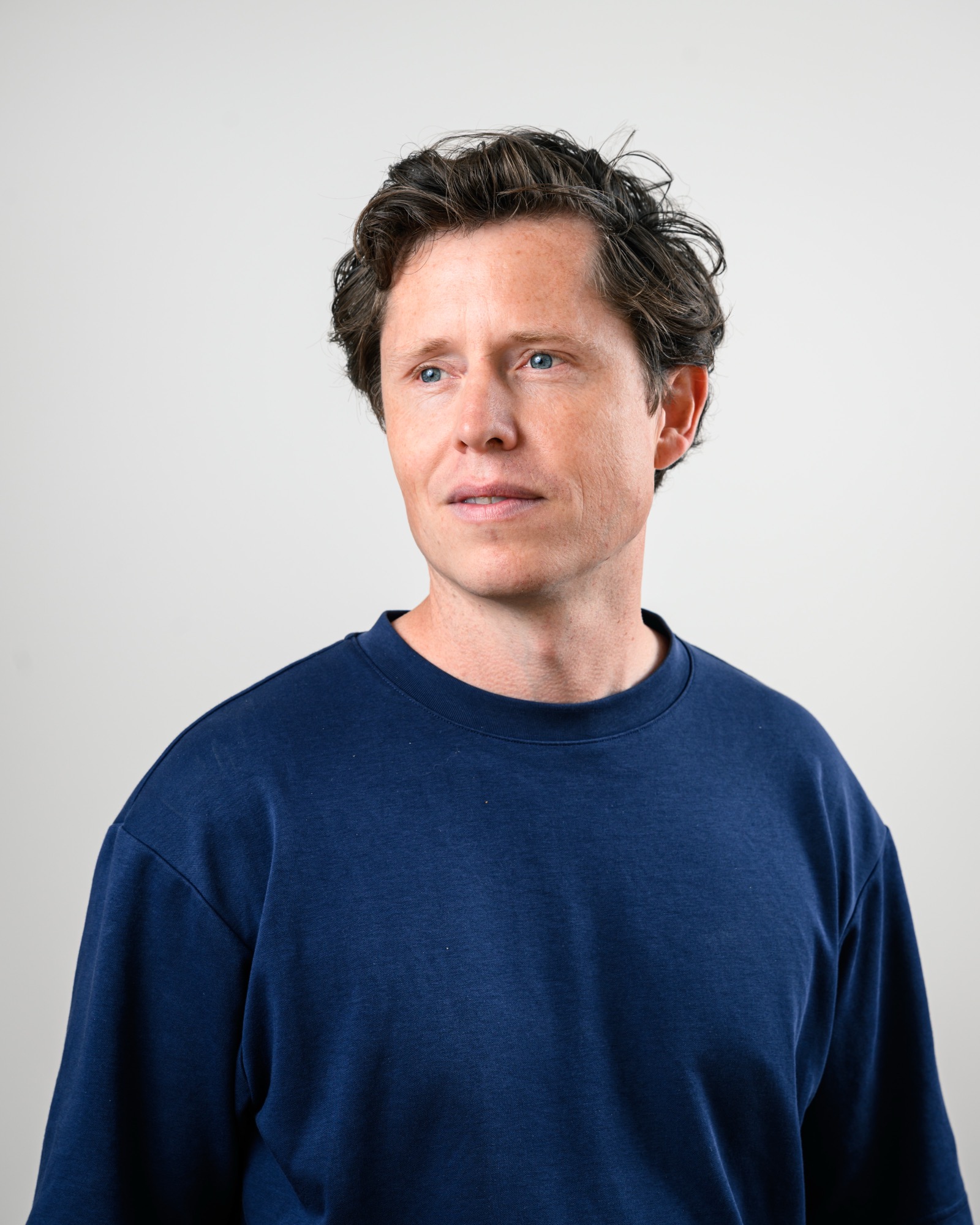 Man with wavy brown hair in a navy blue crew neck t-shirt looking thoughtfully to his left, posed against a neutral light grey background.