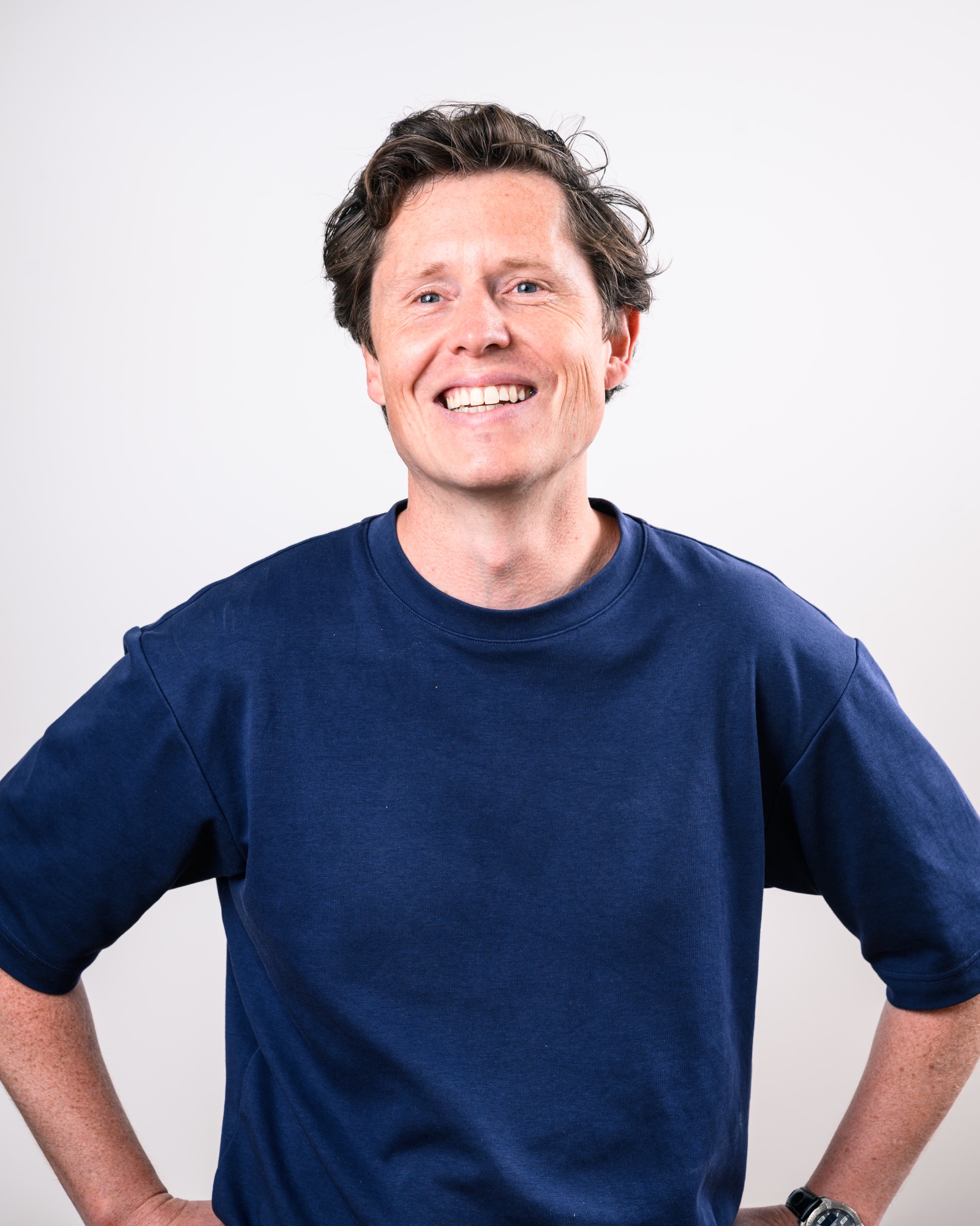 Smiling man with wavy brown hair wearing a navy blue crew neck t-shirt, standing confidently with hands on hips against a light grey background. 