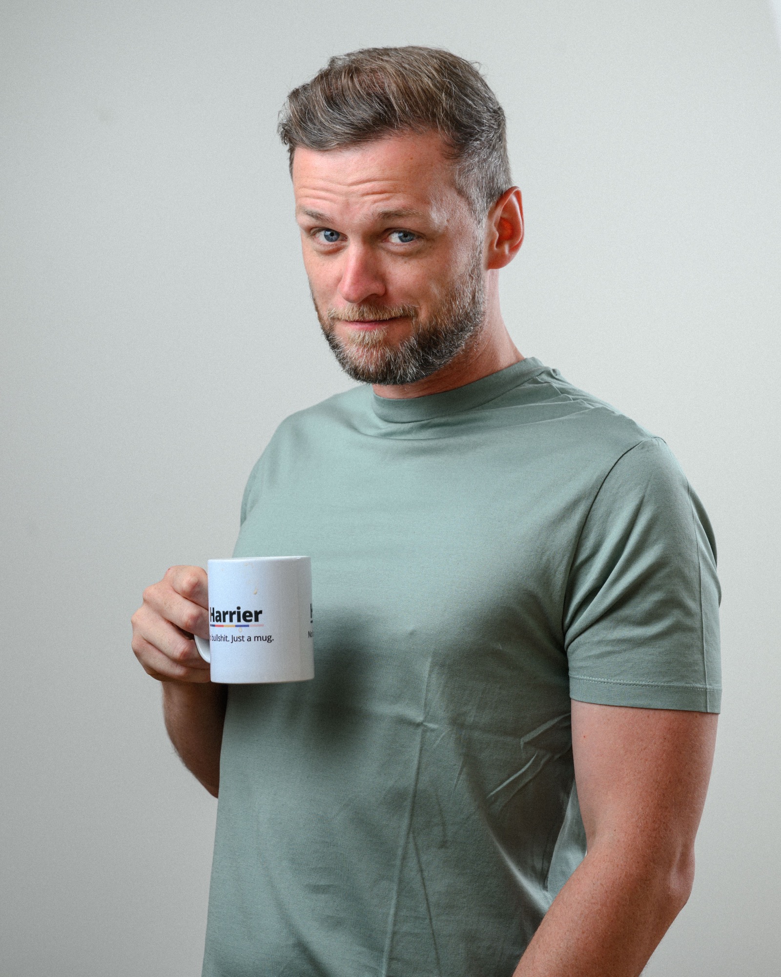 Man with short styled hair and a trimmed beard, wearing a sage green t-shirt, holding a white mug labeled "Harrier" with a playful expression, standing against a light grey background.