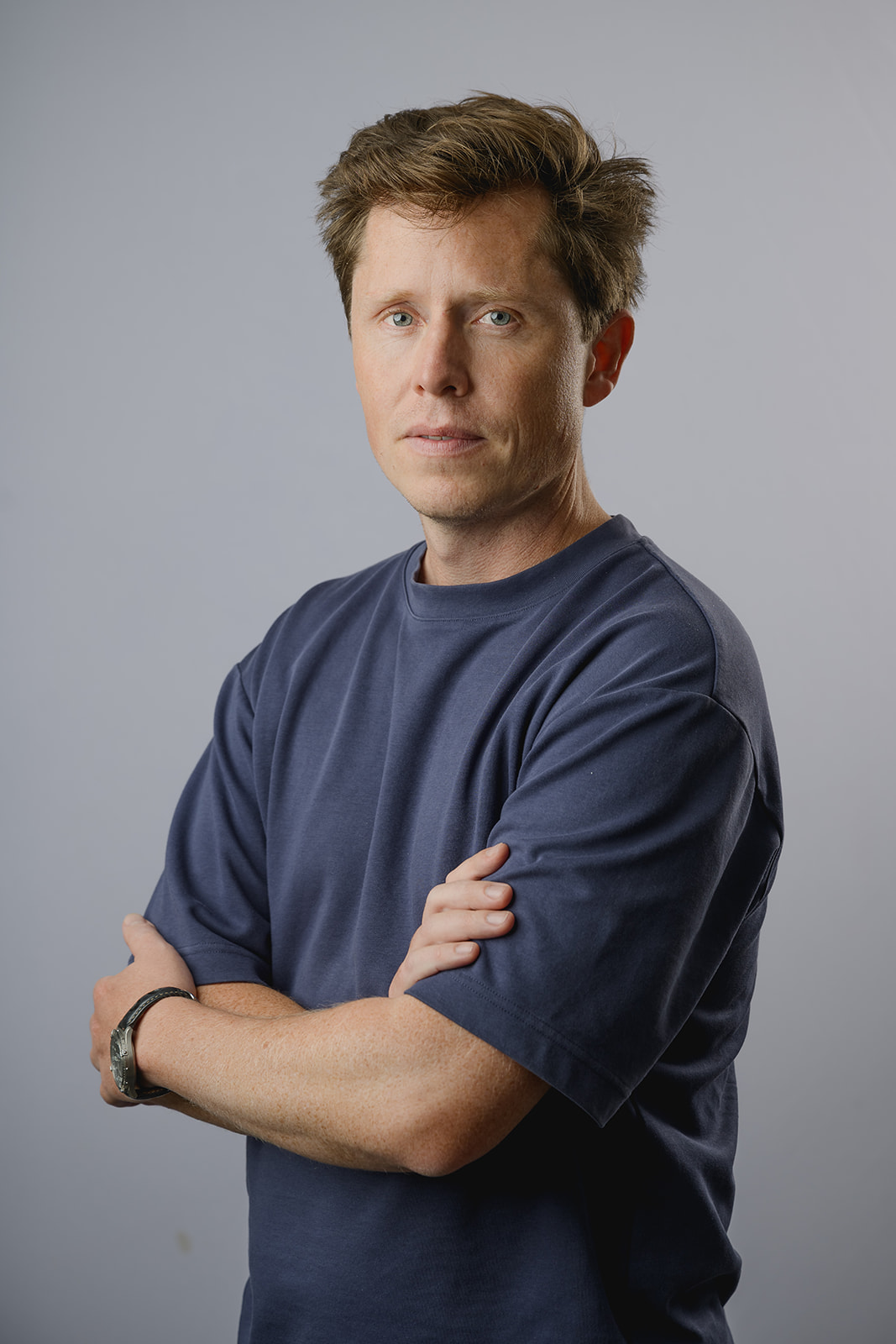 Man with wavy brown hair in a navy blue crew neck t-shirt looking thoughtfully to his left, posed against a neutral light grey background.