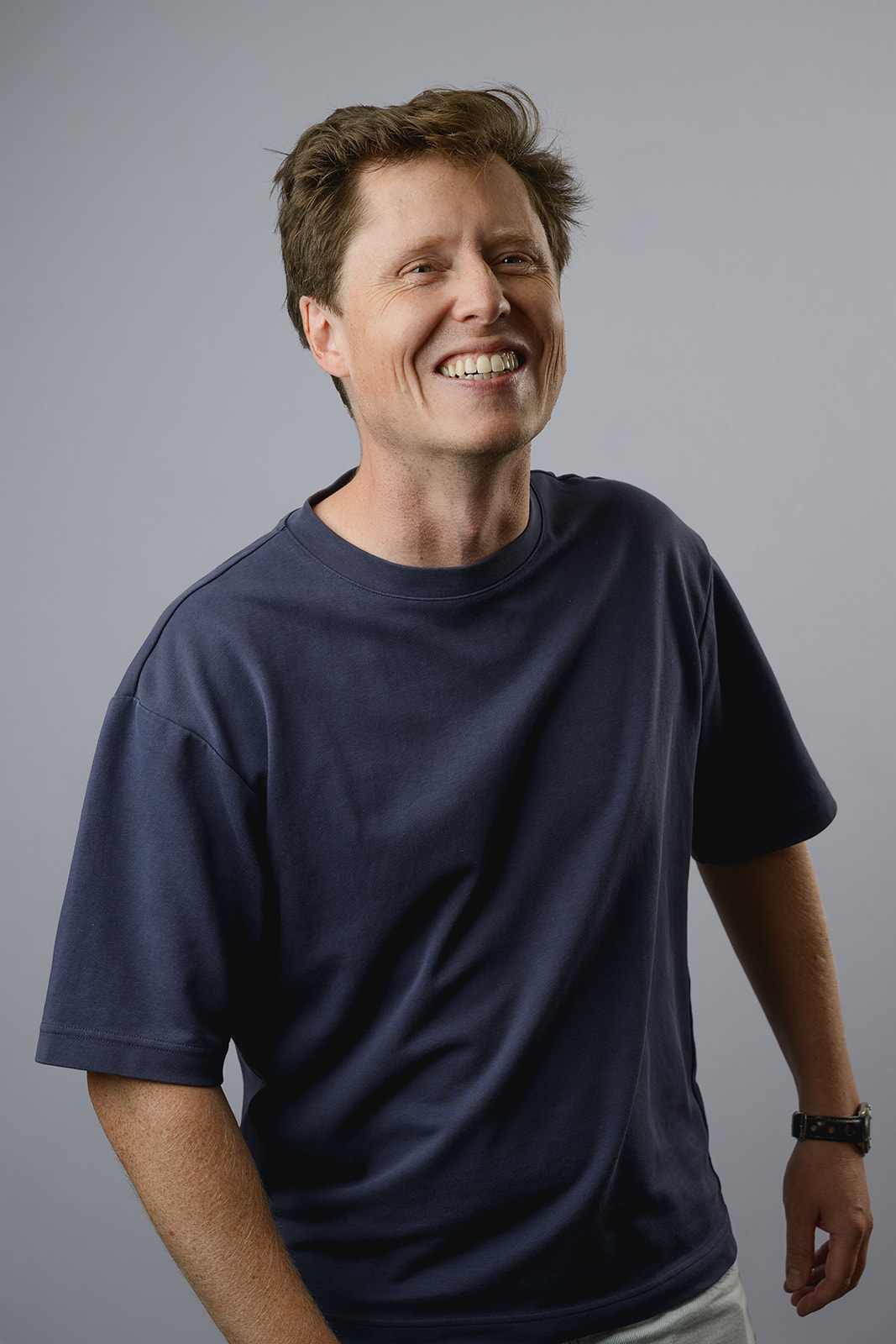 Smiling man with wavy brown hair wearing a navy blue crew neck t-shirt, standing confidently with hands on hips against a light grey background.