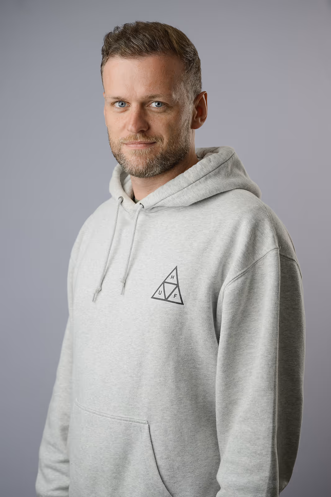 Smiling man with short styled hair and a trimmed beard in a sage green t-shirt, standing casually against a neutral light grey background.