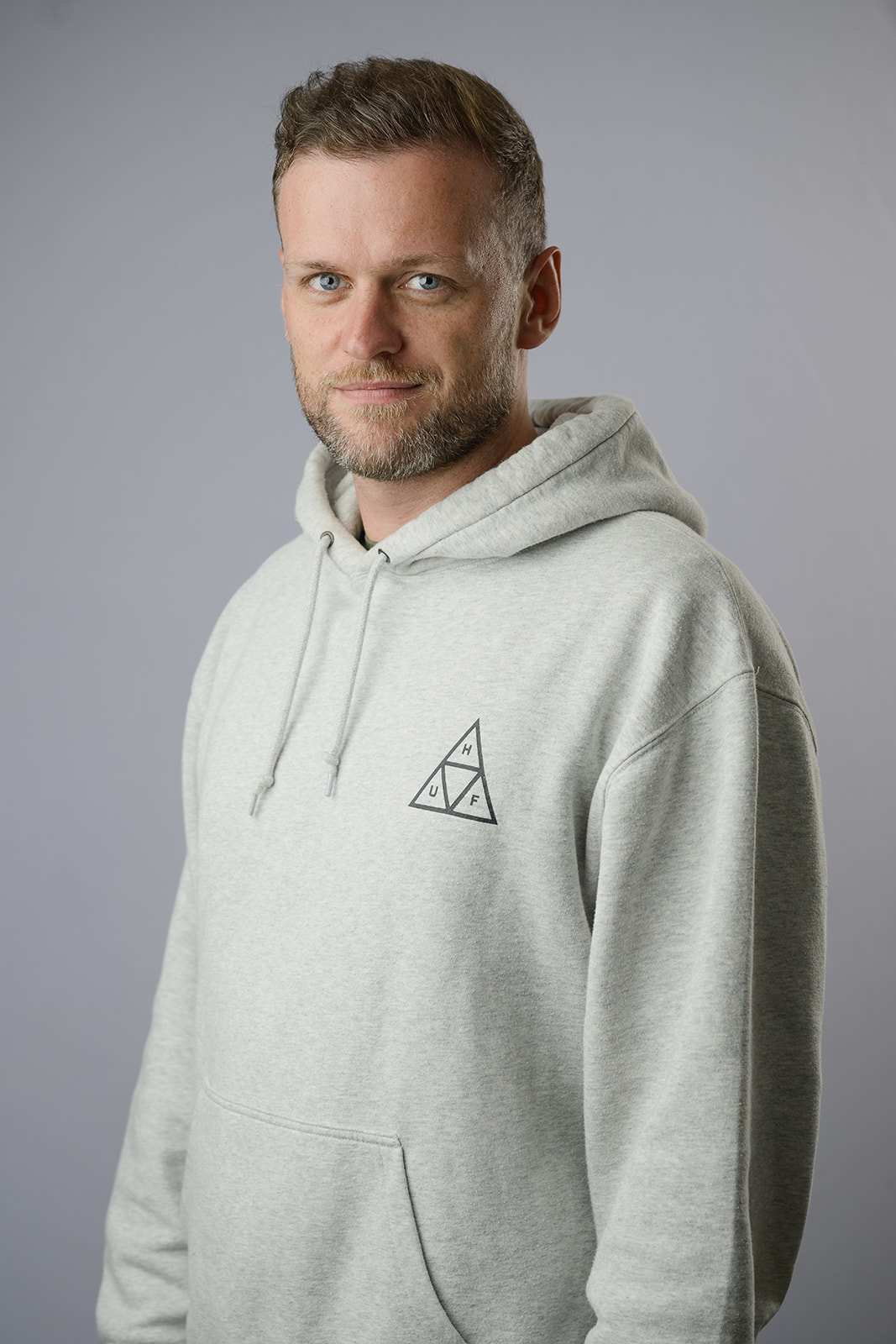 Smiling man with short styled hair and a trimmed beard in a sage green t-shirt, standing casually against a neutral light grey background.